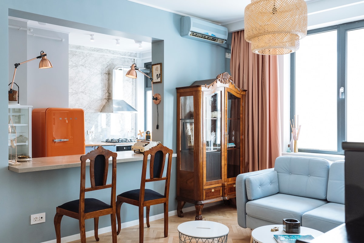 A stylish living area features a light blue sofa, two wooden chairs, and a vintage cabinet. An orange refrigerator adds a pop of color, while a well-equipped kitchen is seen in the background. Natural light enters through large windows adorned with soft curtains.