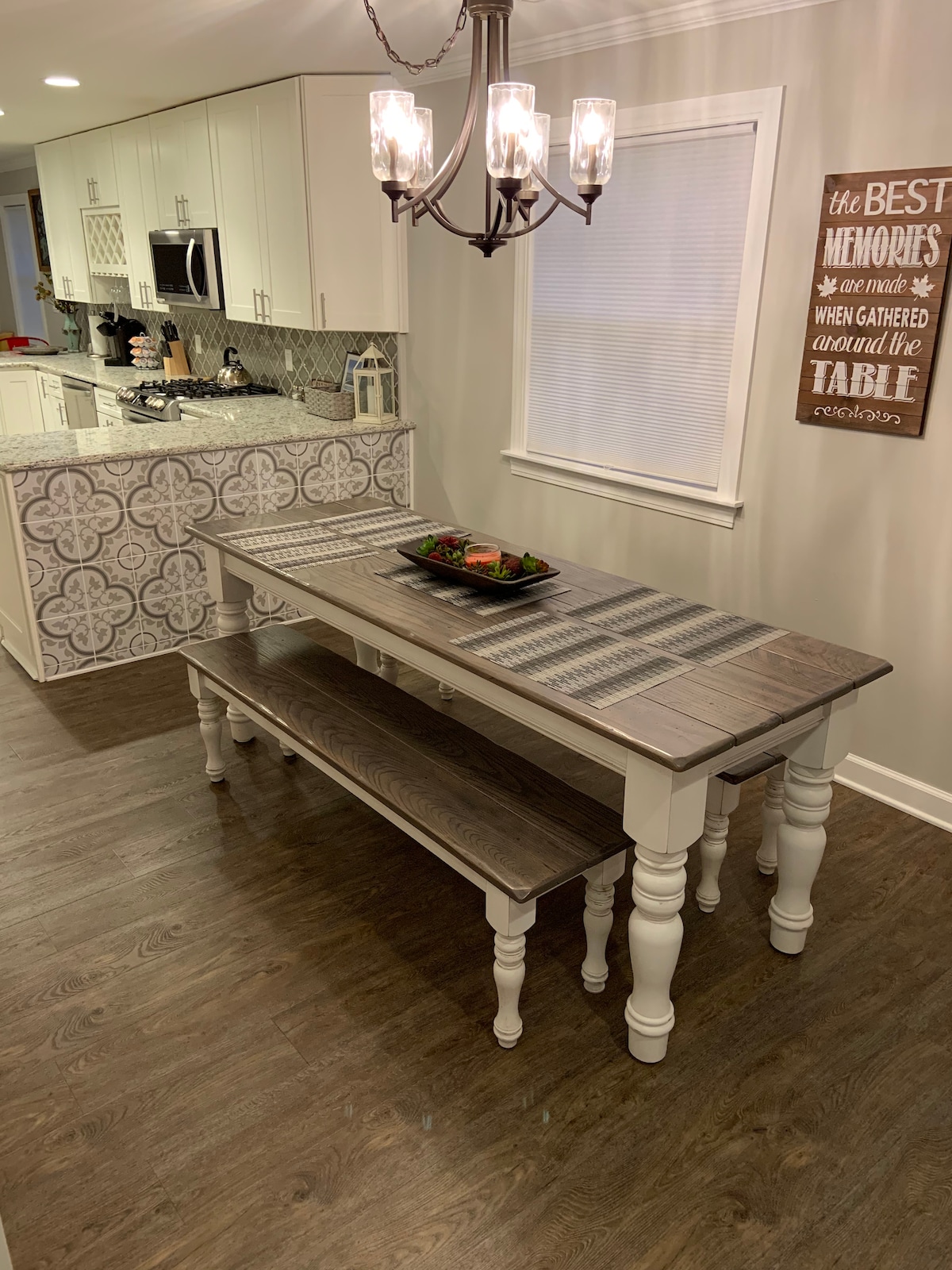 A dining table with a light wooden top is paired with two benches, set against a backdrop of a modern kitchen. A decorative bowl is centered on the table. A stylish light fixture hangs above, and a wall sign offers a warm message about memories made around the table.