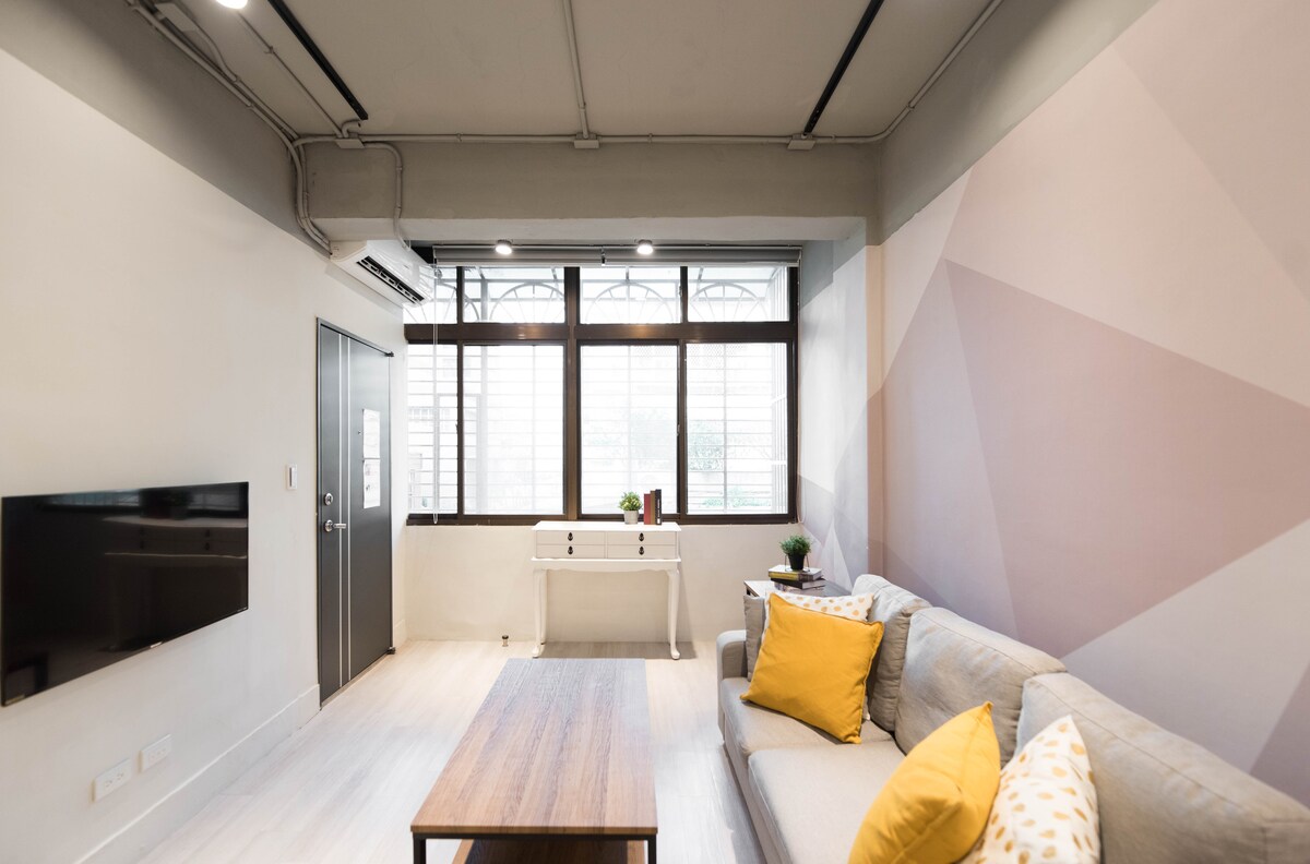 A modern living area is presented with large windows allowing ample natural light. A simple gray sofa with yellow accent pillows sits across from a wall-mounted TV. A minimalist wooden coffee table is positioned in the center, complemented by a small decorative table featuring greenery.