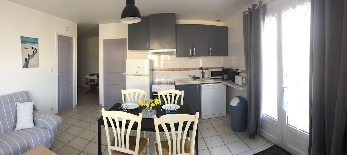 A modern kitchen and dining area are visible, featuring a table set for four with a vase of flowers. The kitchen is equipped with grey cabinetry, a refrigerator, and various appliances including a microwave and toasting machine. Natural light enters through the glass doors leading to the outdoor space.
