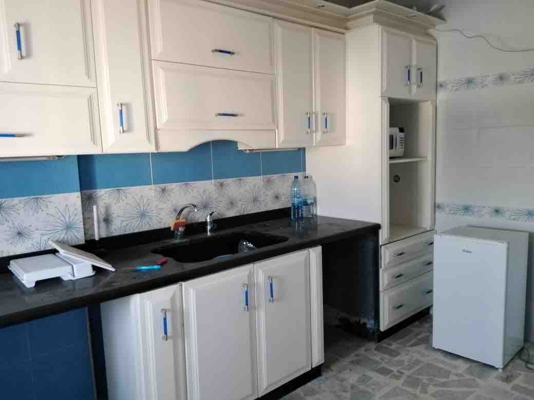 The kitchen features white cabinets with blue accents, complemented by a dark countertop. A double sink is present beneath a window, and a small refrigerator is located nearby. Additional appliances like a microwave are integrated into the cabinetry, with ample storage provided by upper and lower cabinets.