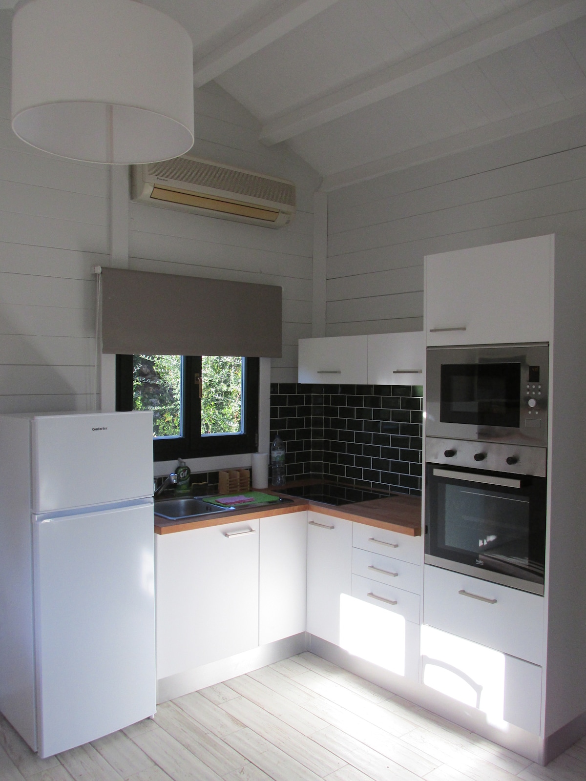 A modern kitchen area is equipped with essential appliances, including a refrigerator and an oven. The cabinets feature a combination of light and dark colors, with dark green tiles on the backsplash. Natural light filters in through a nearby window, illuminating the open space.