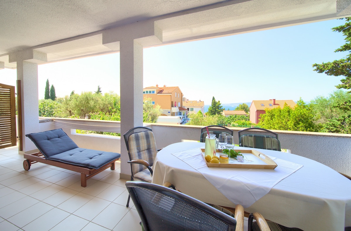 An outdoor terrace with a dining table set for six is visible, surrounded by comfortable chairs. A lounge chair is positioned nearby for relaxation. Scenic views of greenery and distant rooftops, with the horizon hinting at the sea, can be seen in the background.