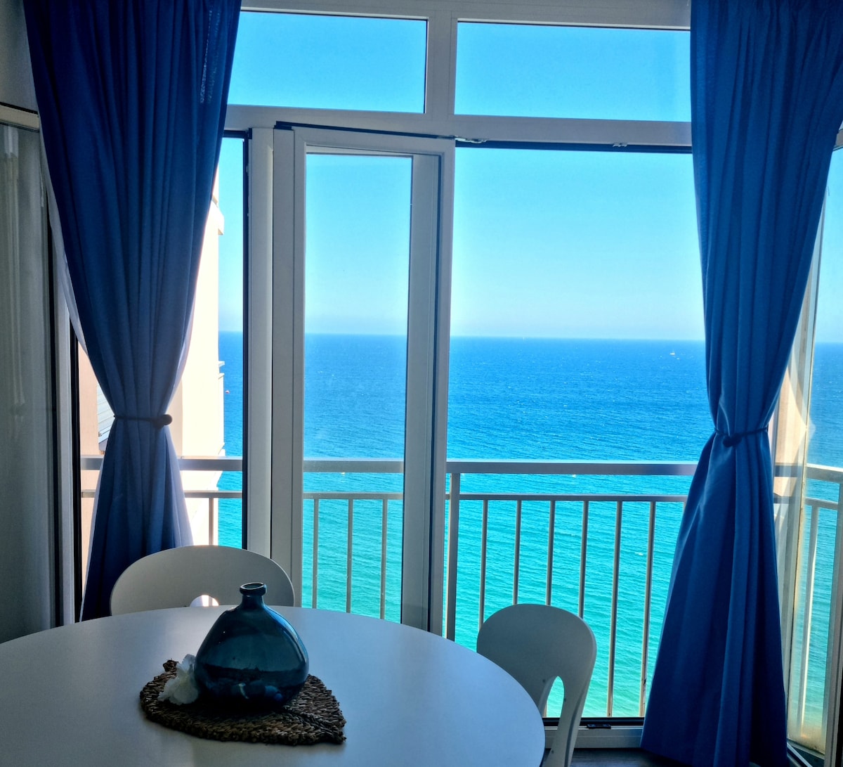 Large windows offer expansive views of the Mediterranean Sea, with blue curtains framing the scene. A dining table is set near the glass doors, accompanied by a decorative bowl at its center. The serene ocean reflects shades of blue under a clear sky.