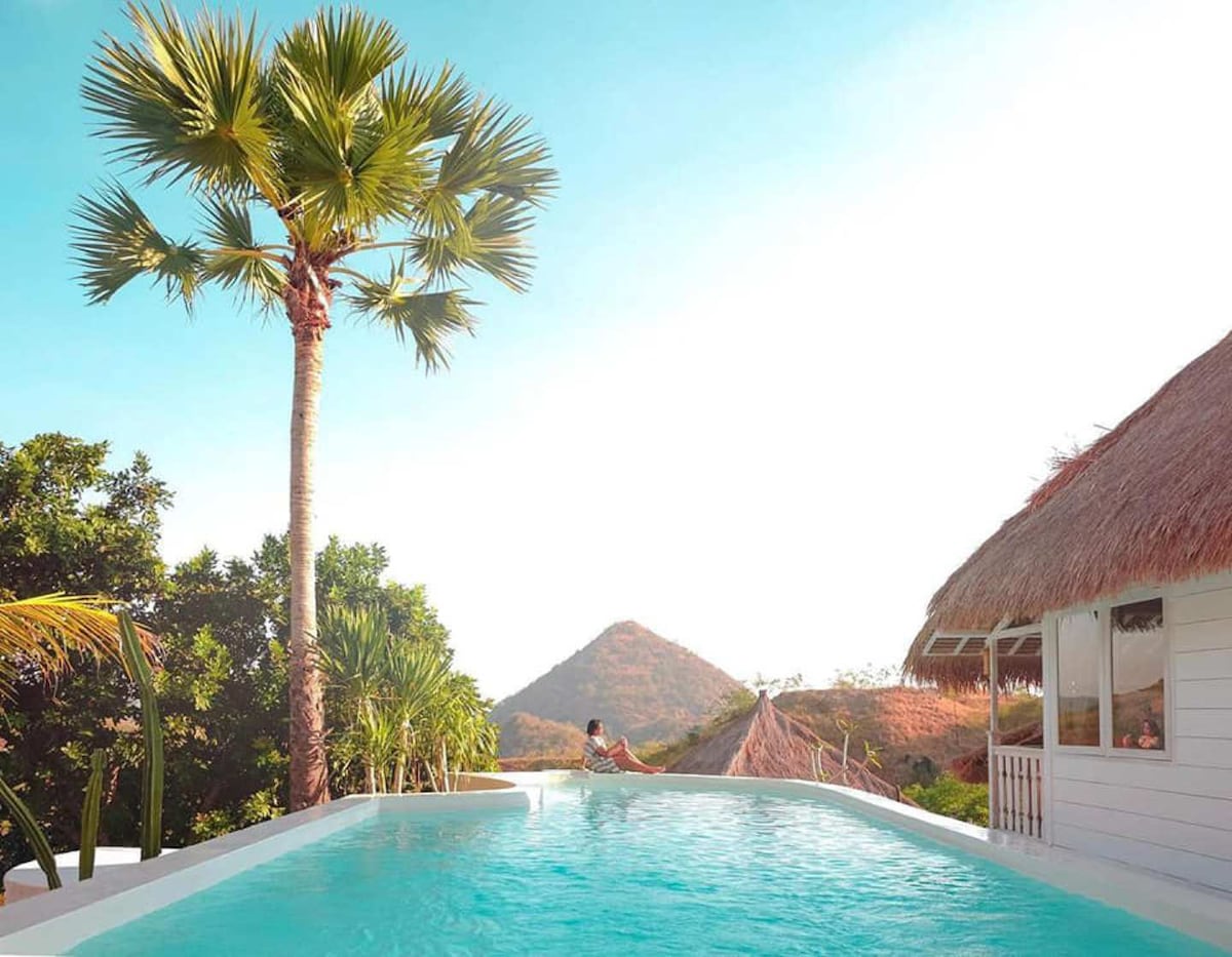 An expansive pool with clear blue water is surrounded by lush greenery and palm trees. A thatched-roof bungalow is positioned nearby, while a gentle hill rises in the background, under a bright blue sky.