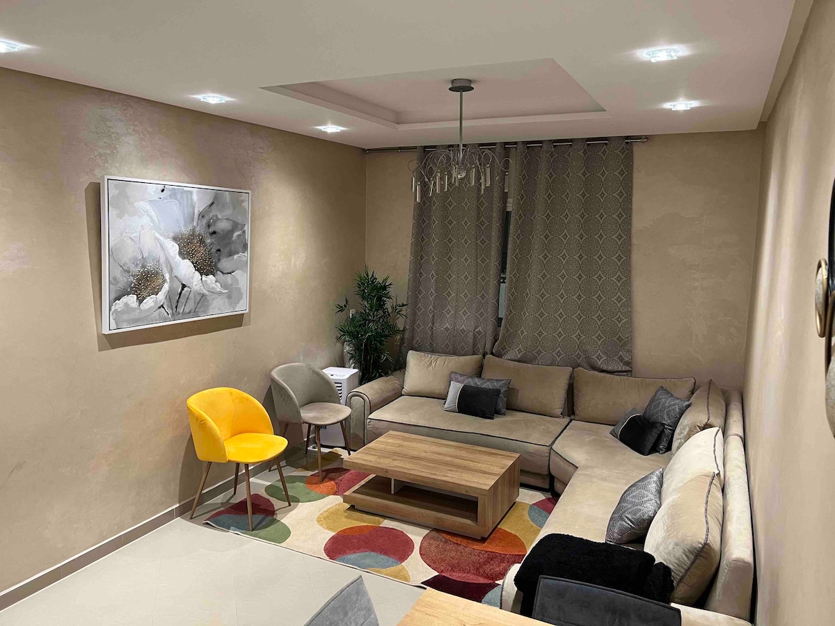 A contemporary living area features a large, U-shaped sofa with a mix of neutral tones and colorful accent pillows. A wooden coffee table sits at the center, surrounded by a variety of seating options, including a bright yellow chair. Soft, ambient lighting is provided by recessed fixtures.