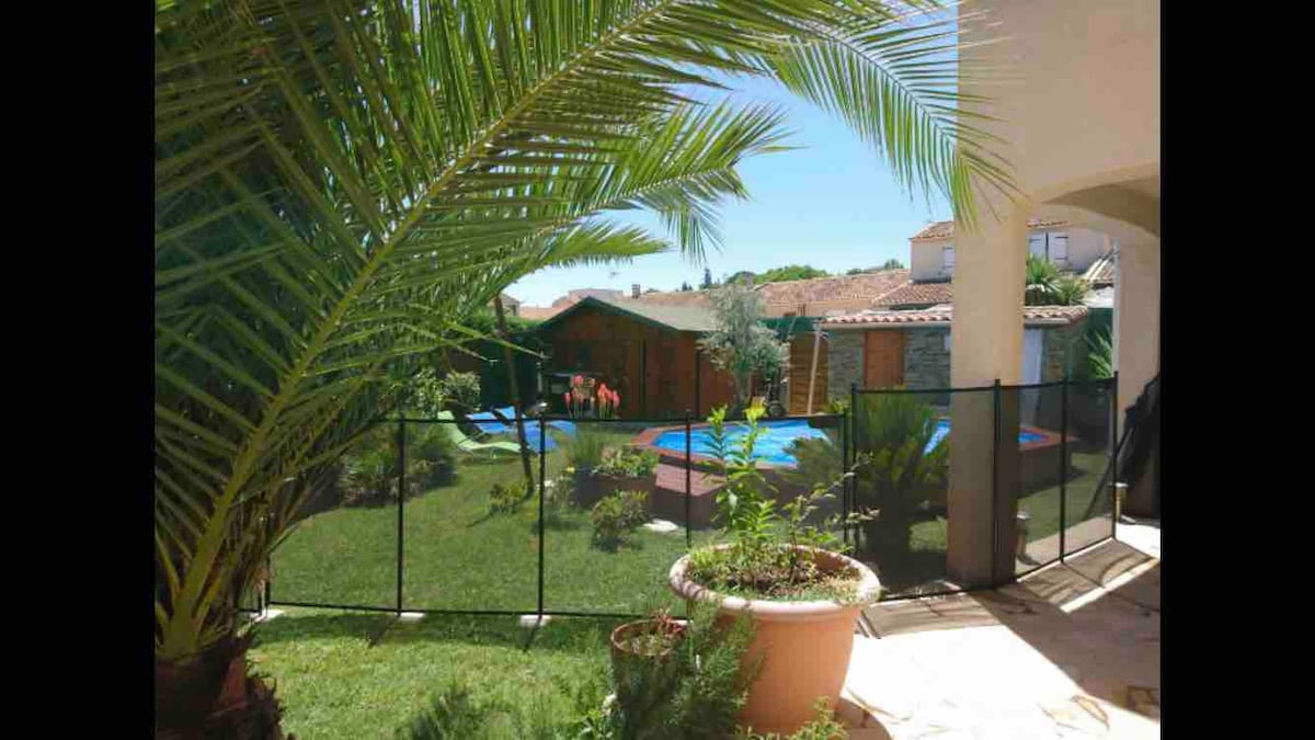 A lush garden area is visible, framed by tall palm leaves. A fenced pool area is centrally located, surrounded by greenery and colorful flowers. In the background, wooden cabins are set against a clear blue sky, contributing to a serene outdoor environment.