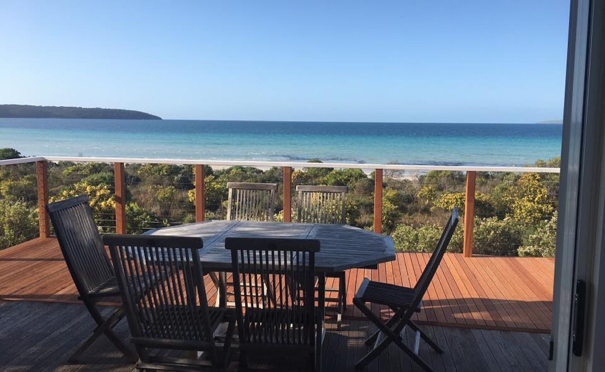 A spacious outdoor deck is furnished with a round wooden table and six wooden chairs, offering a direct view of the tranquil blue ocean and lush coastal vegetation. The sea's gentle waves can be seen lapping at the shore, enhancing the serene atmosphere.