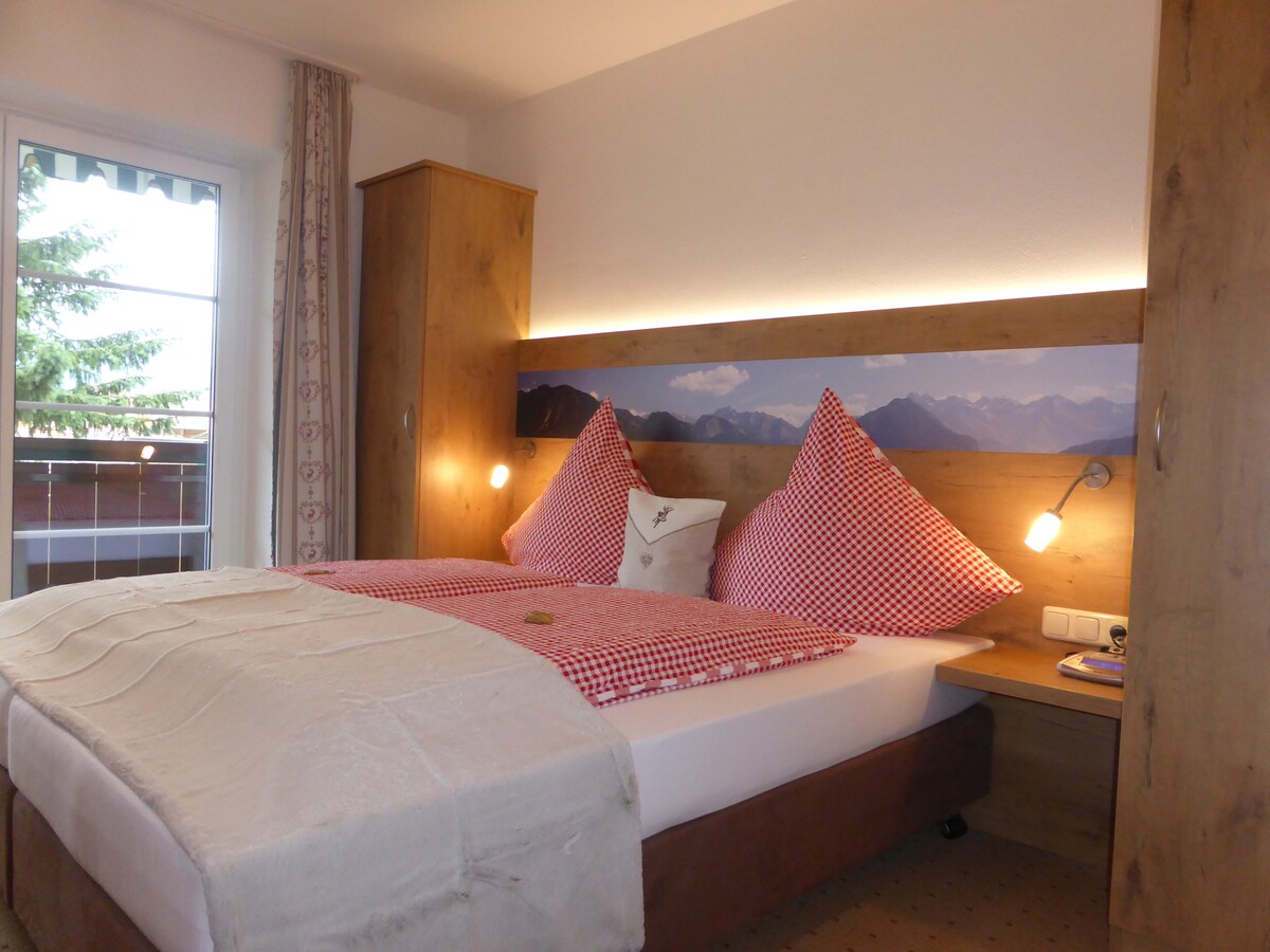 A cozy bedroom features a spacious bed adorned with red and white checked pillows and a soft, light blanket. Soft lighting is provided by wall-mounted lamps, and a scenic mural of mountains enhances the calming environment. A window offers natural light, complemented by curtains.