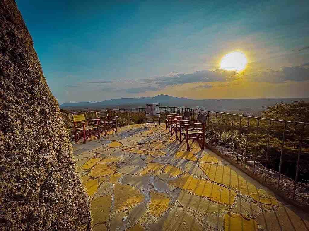 A spacious terrace is depicted with wooden chairs arranged to face the horizon, where a sunset casts a warm glow. The stone-paved floor is visible, leading to a panoramic view of distant hills and valleys under a gently illuminated sky.