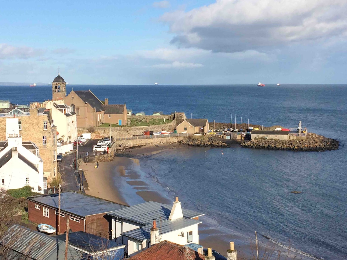 The image captures a coastal view featuring a sandy beach alongside a quaint harbor. A mix of residential and commercial buildings is seen, with boats docked at the shore. The horizon displays a calm sea under a partly cloudy sky, creating a serene seaside atmosphere.