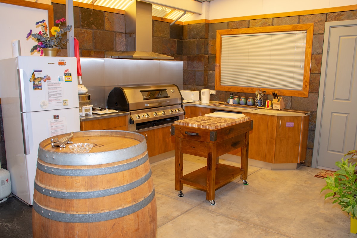 A spacious kitchen area is equipped with modern appliances, including a large stainless steel gas grill. A wooden preparation table and a wine barrel add character to the space. Ample countertops are complemented by wooden cabinetry and a large window providing natural light.