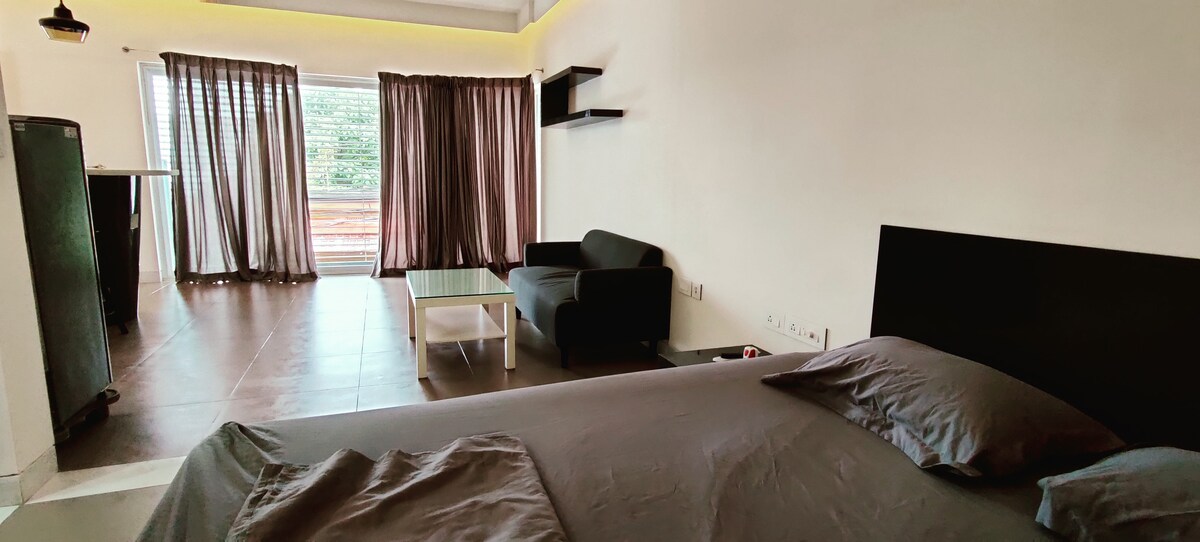 A spacious studio apartment interior is visible, featuring a large bed with dark bedding. A cozy seating area includes a dark sofa and a glass coffee table. Natural light enters through large windows, enhanced by sheer curtains, creating an open and inviting atmosphere.