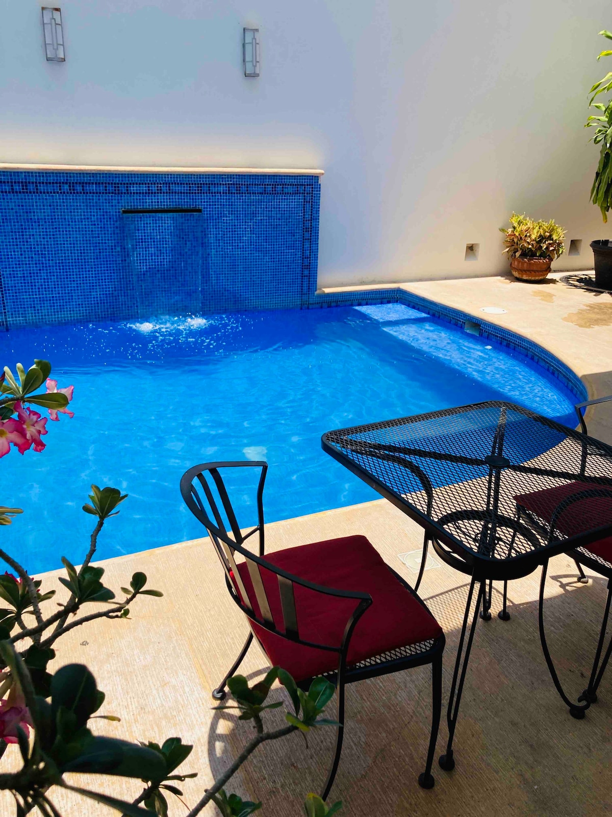 A private pool features vibrant blue tiles with a gentle waterfall, surrounded by a smooth stone deck. A small table and two chairs in black metal are positioned beside the pool, complemented by lush greenery and flowering plants nearby.