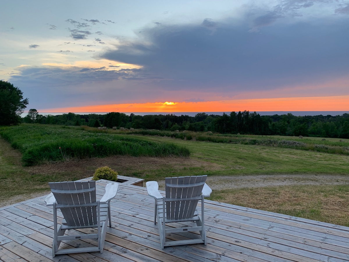 Two white Adirondack chairs are positioned on a wooden deck, facing a colorful sunset over the expansive landscape. The sky features shades of orange and purple, while grassy fields stretch towards the horizon, creating a serene outdoor setting.