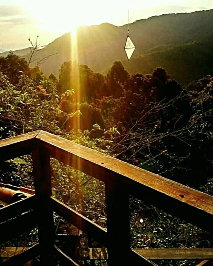 Golden sunlight streams over the mountain ranges, illuminating lush greenery. A wooden railing creates a warm frame, with the view of trees extending into the distance. A prism catches the light, adding a subtle sparkle to the serene landscape.