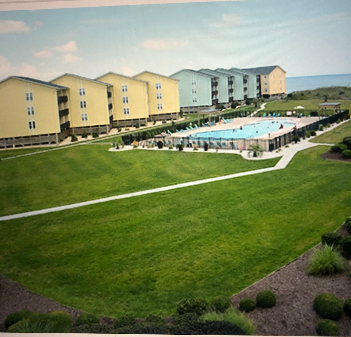 The image presents a spacious outdoor area featuring a well-maintained swimming pool surrounded by neat landscaping. Several multi-story buildings are visible in pastel colors, with expansive green lawns and walkways leading to the pool area, all set against a backdrop of the ocean.