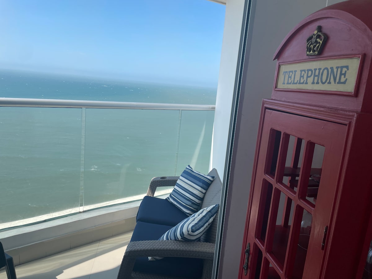 A balcony offers a stunning view of the ocean, with a comfortable seating area featuring striped cushions. A red telephone booth adds a unique design element to the space, enhancing the overall aesthetic of this coastal retreat.