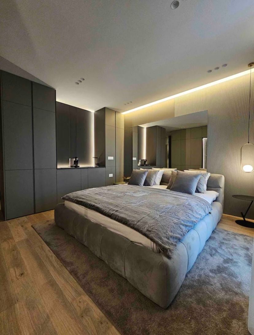A spacious bedroom features a plush bed with soft bedding and several pillows. Modern cabinetry lines the walls, while warm lighting enhances the sleek design. A small side table and an area rug add comfort to the overall aesthetic.