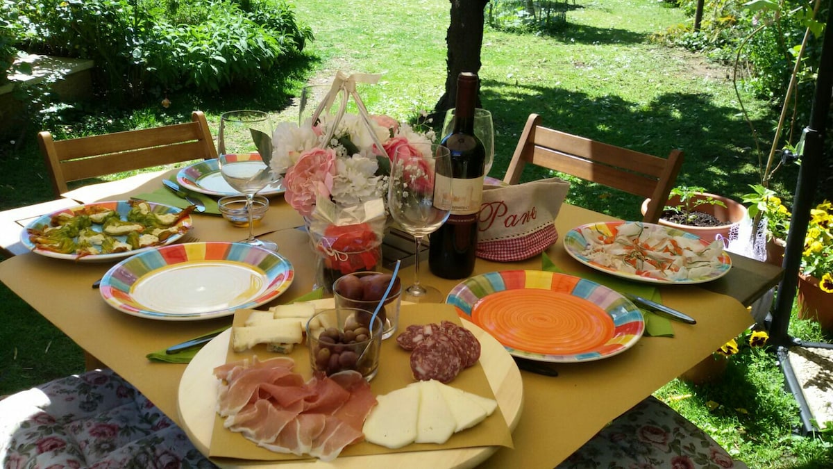 A wooden table is set in a green garden, featuring colorful plates with various food items including cured meats, cheeses, olives, and a selection of appetizers. Glasses are present, along with a bottle of wine. Surrounding the table, vibrant flowers and greenery create a relaxed outdoor dining scene.