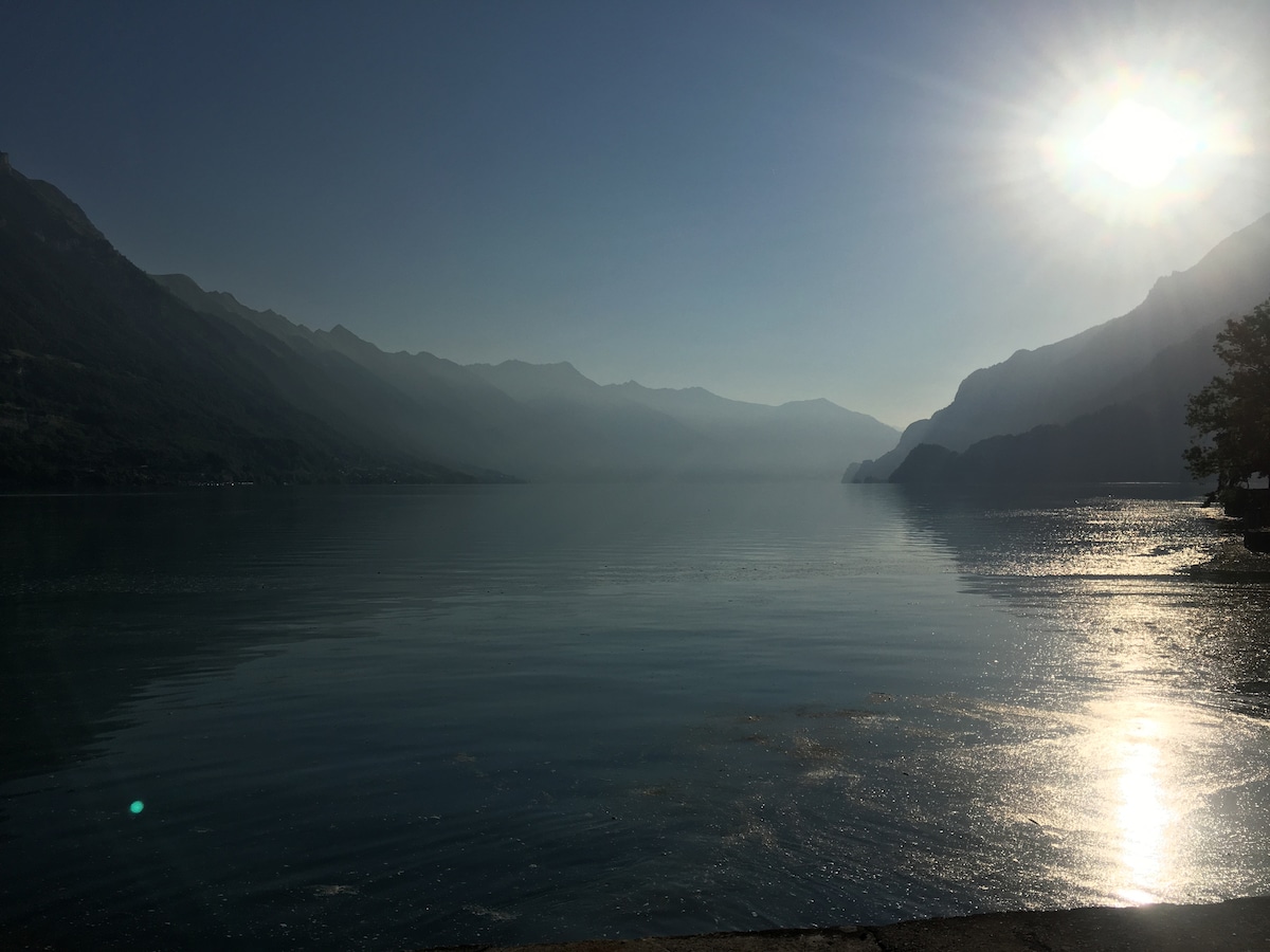 A calm lake is viewed in the early morning light, reflecting the surrounding mountains. The sun hovers low in the sky, casting a gentle glow over the water, while a soft mist envelops the distant landscape.