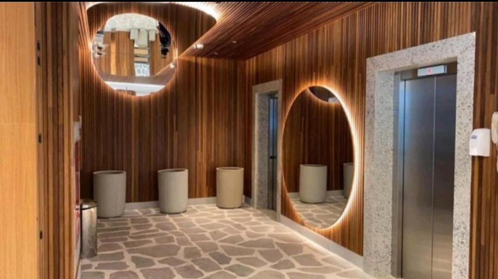 The entryway features warm wooden paneling complemented by circular mirrors that enhance the light ambiance. A smooth stone floor is visible, leading to an elevator with a metallic finish, and two sleek, gray planters add a modern touch to the space.