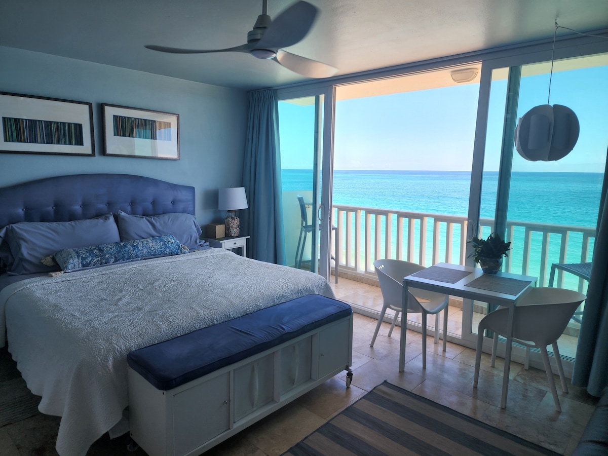 A studio apartment features a spacious bed dressed in white linens and a blue decorative throw. A dining table with chairs overlooks a panoramic ocean view through large sliding glass doors. Soft blue walls complement framed artwork and a ceiling fan enhances the airy feel.