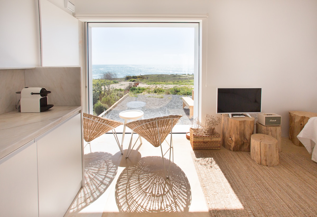 A bright living area features large glass doors that open to a view of the sea. Two wicker chairs are positioned on a textured area rug, while a small television and decorative wooden elements add to the minimalistic decor. Natural light floods the space.