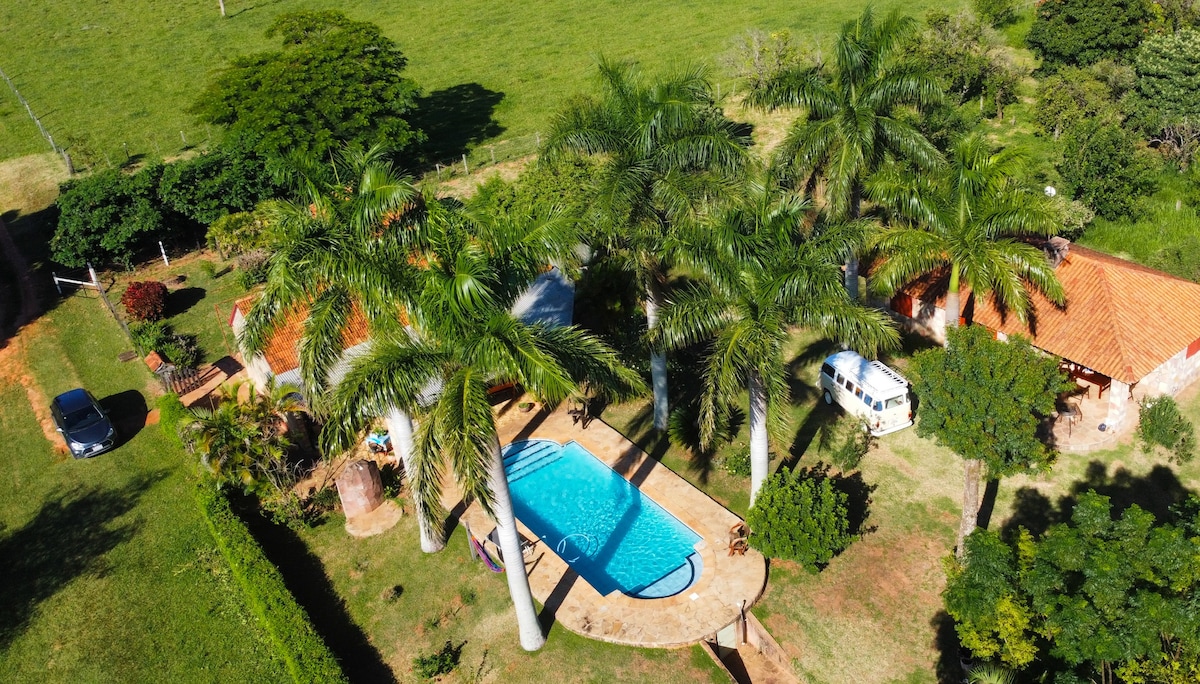 An aerial view of the property showcases a bright blue swimming pool surrounded by tall palm trees. The lush green landscape provides a peaceful backdrop, while a charming house with a red-tiled roof is visible nearby, enhancing the natural tranquility of the setting.