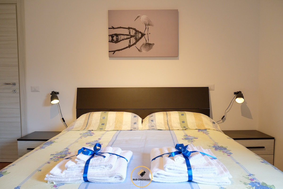 A serene bedroom features a spacious bed with a beige and blue-striped coverlet. Two neatly folded towels tied with blue ribbons are placed at the foot of the bed. Wall-mounted reading lights are located on either side, and a framed black-and-white photograph of herons adorns the wall above.