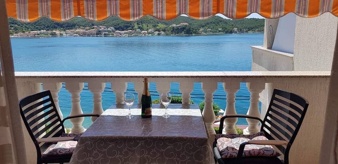A balcony overlooks the tranquil blue waters, featuring a table set for two with a bottle of sparkling wine and glasses. Two black chairs flank the table, and an orange and white striped awning provides shade from the sun.