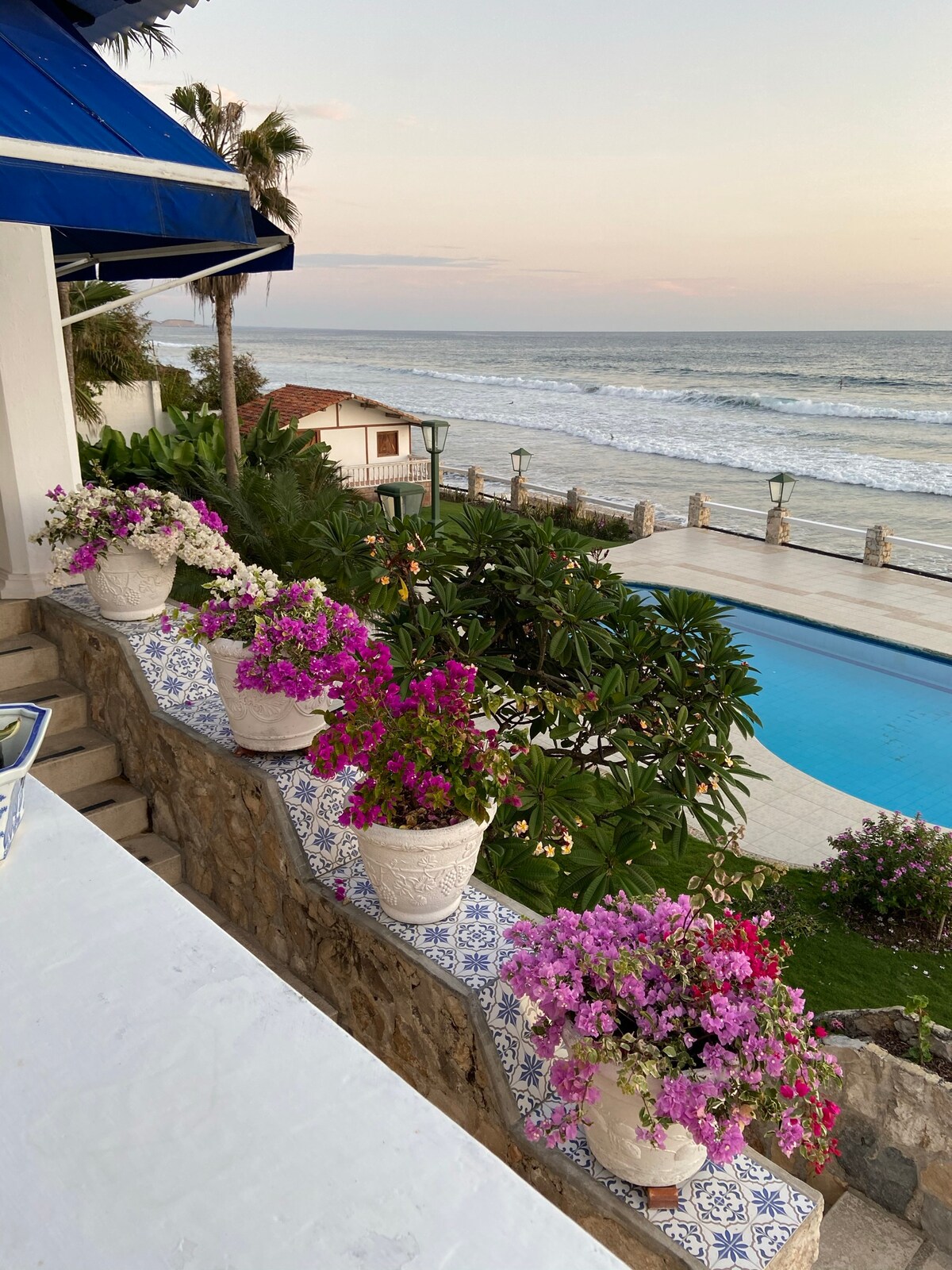 Colorful flower pots line a stone wall, their vibrant blooms contrasting with the serene blue of the swimming pool and the ocean waves beyond. The horizon showcases a gentle sunset, enhancing the tranquil ambiance of the coastal landscape.