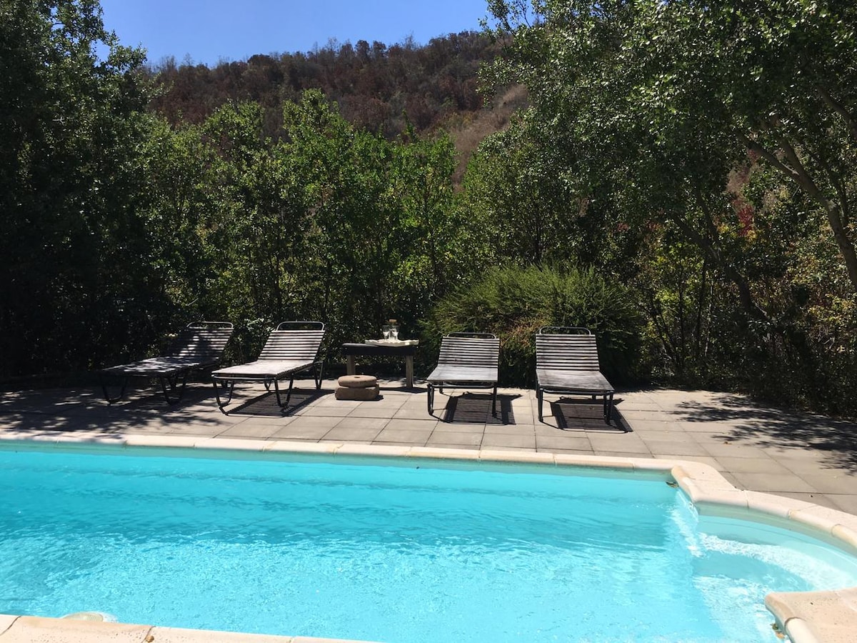 A refreshing swimming pool is surrounded by a spacious patio. Several lounge chairs are arranged nearby, set against a backdrop of lush greenery and gently rolling hills. Sunlight reflects off the clear water, creating a relaxed and inviting outdoor space.
