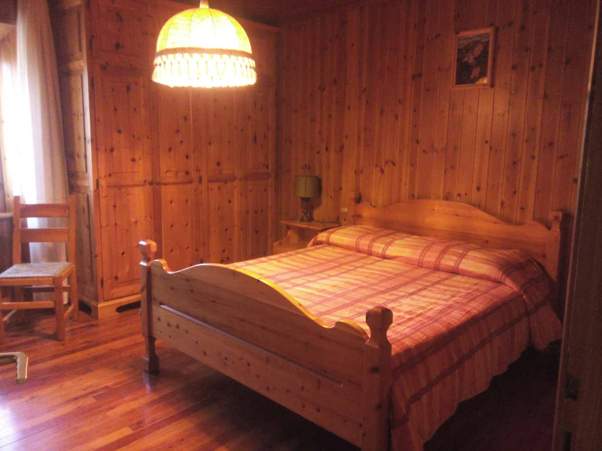 A warm-toned bedroom features wooden walls and flooring, creating a cozy environment. A spacious bed with a plaid coverlet is centered in the room, complemented by a simple chair and a bedside lamp. Natural light filters through a window, enhancing the inviting feel.