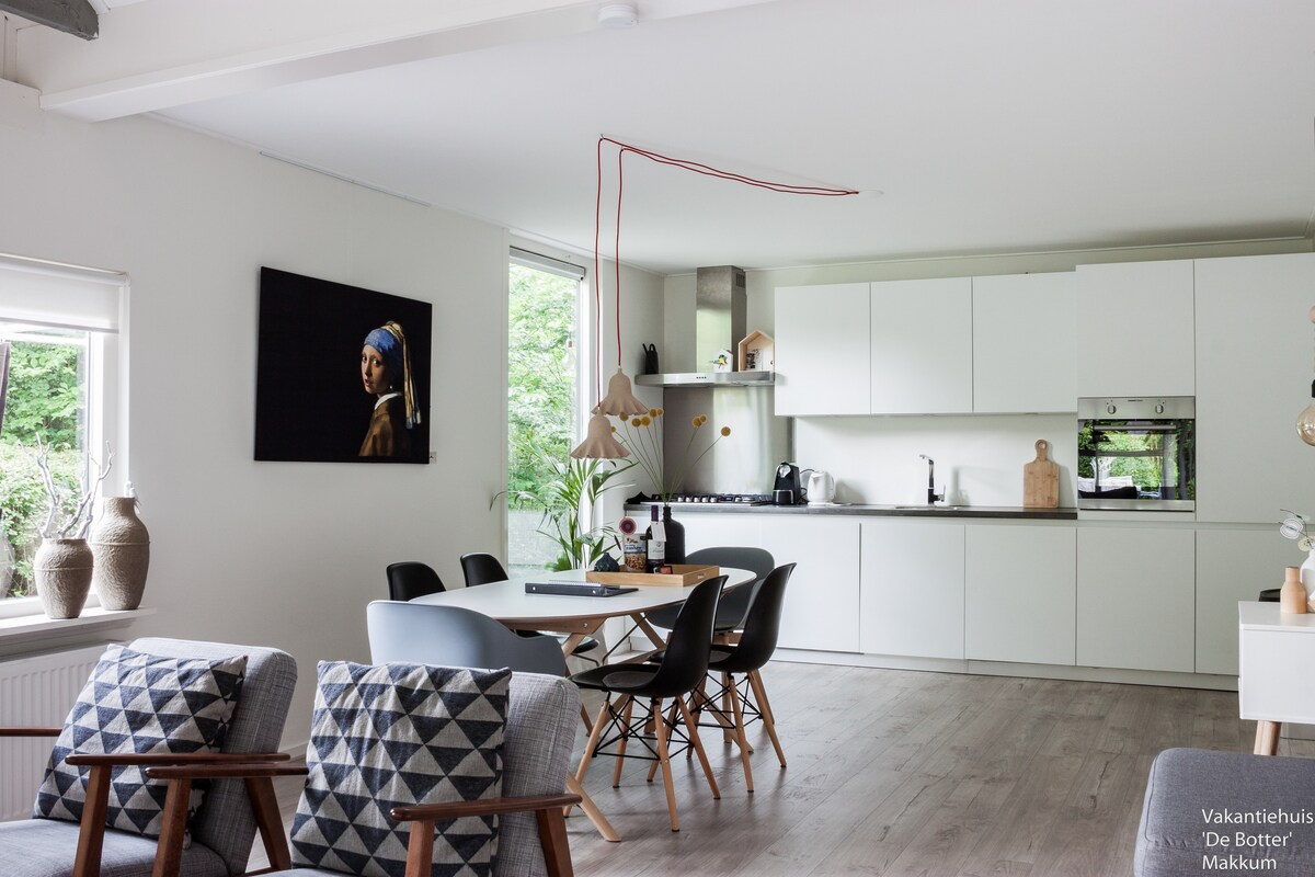 A modern kitchen area is visible, featuring white cabinetry and sleek appliances, including an oven and a stovetop. A dining table with dark chairs is positioned in front of the kitchen, while a contemporary artwork is displayed on the wall, creating an inviting dining space.