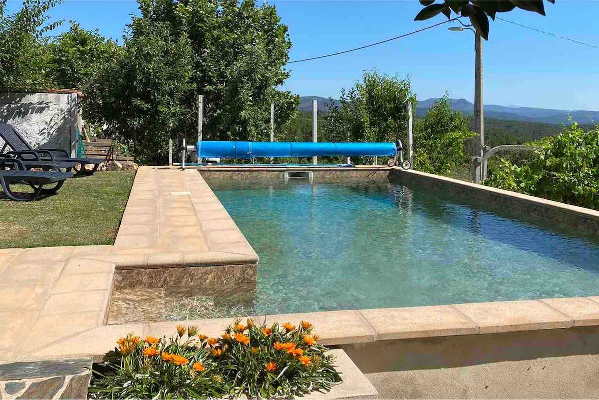 A heated outdoor salt pool is surrounded by natural greenery, offering scenic views of the mountains. Sun loungers are positioned nearby for relaxation, and a blue pool cover is rolled up at one end. Vibrant flowers bloom at the edge of the pool area.