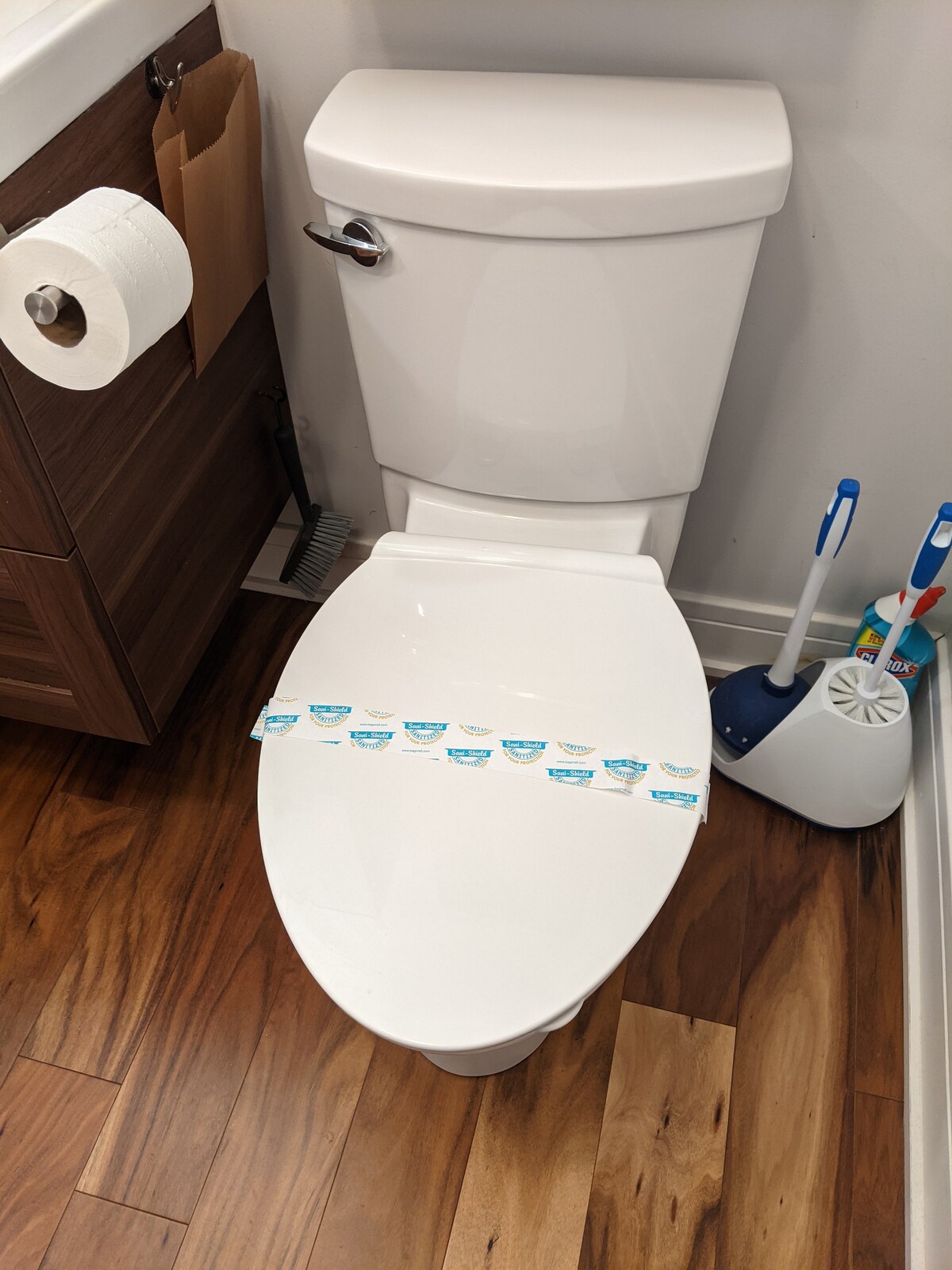 The bathroom features a clean, modern toilet with a fresh seal for guest assurance. A roll of toilet paper is placed conveniently nearby, along with a toilet brush and holder. The flooring is rich brown, enhancing the overall warmth of the space.