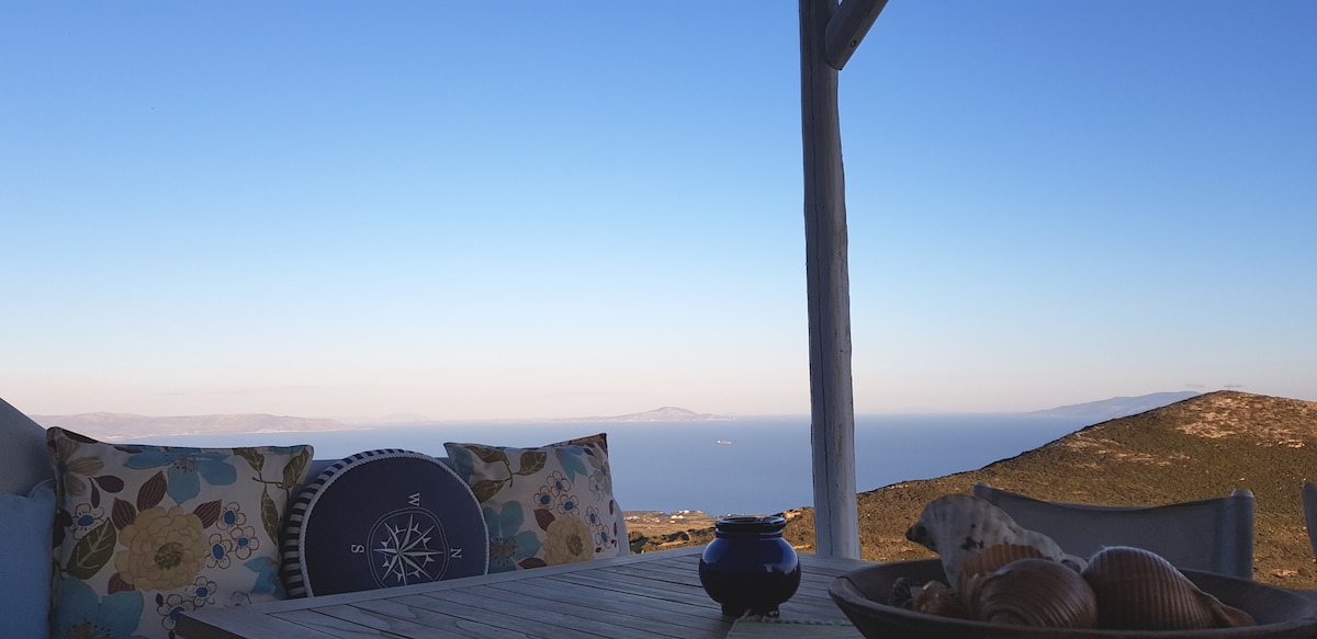 An outdoor seating area is presented with floral cushions arranged on a wooden bench, overlooking a vast blue sea and distant islands. A decorative bowl filled with shells is centered on the table, complementing the tranquil surroundings under a clear sky.