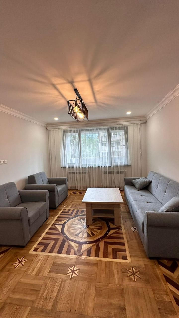 A living area features two grey sofas and a matching armchair, arranged around a light wooden coffee table. Natural light flows in through a large window, complemented by sheer curtains. The elegant parquet flooring showcases an intricate geometric pattern.