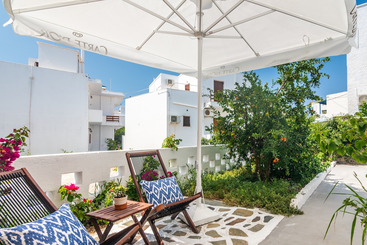 An outdoor seating area is shown, featuring two lounge chairs with patterned cushions and a shaded umbrella. Surrounding greenery includes flowering plants and a fruit tree, creating a calm, private space. The pathway is composed of decorative stones, leading through the inviting garden.