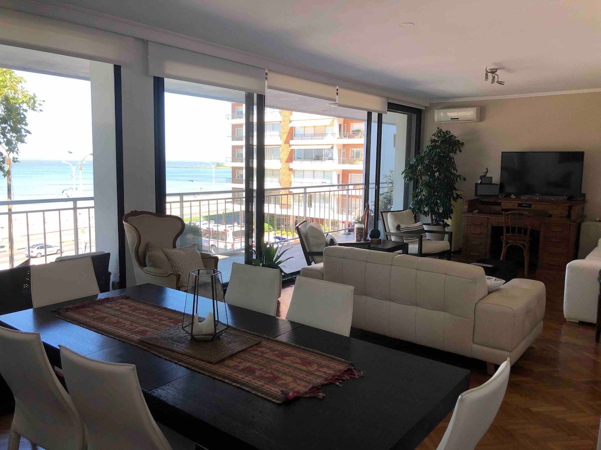 An open living space features large windows offering views of the ocean. A dark dining table is surrounded by bright chairs, while a light-colored sofa and armchairs create a seating area. A decorative table centerpiece adds a subtle detail, complemented by greenery from a nearby plant.