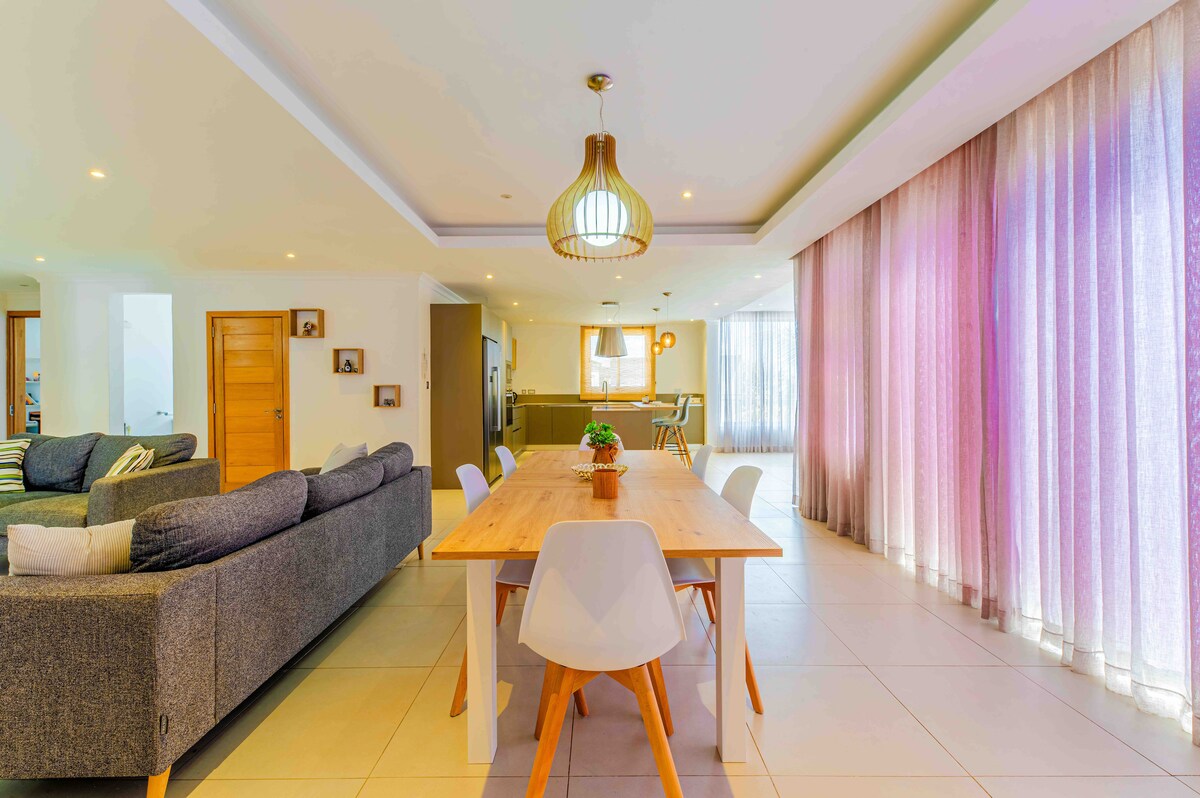 A spacious dining area is illuminated with natural light from large windows adorned with soft curtains. A wooden dining table is surrounded by white chairs, with a light fixture hanging above. In the background, a cozy living space is visible, featuring comfortable seating.