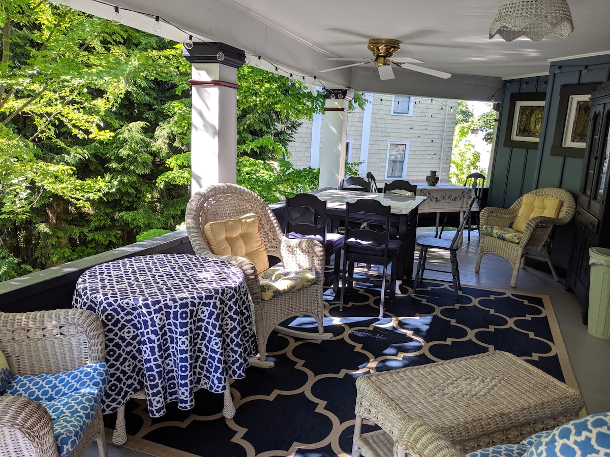 A spacious porch is furnished with wicker seating, including two armchairs and a small table, creating a cozy atmosphere. A large dining table with chairs is positioned at the back, while a patterned rug covers the floor, adding texture to the outdoor space surrounded by greenery.