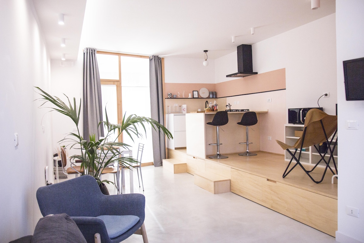A modern studio layout features an elevated area with a cozy kitchen and dining space. Light-colored walls provide a bright atmosphere, complemented by a green plant. A sleek chair and a gray sofa add comfort, while large windows allow natural light to illuminate the area.