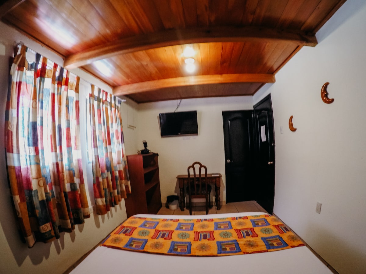 A brightly lit room features a spacious bed covered with a patterned blanket. Colorful curtains frame a window, allowing natural light to enhance the space. A wooden table with a chair and a wall-mounted television are also present, contributing to the room's functionality.