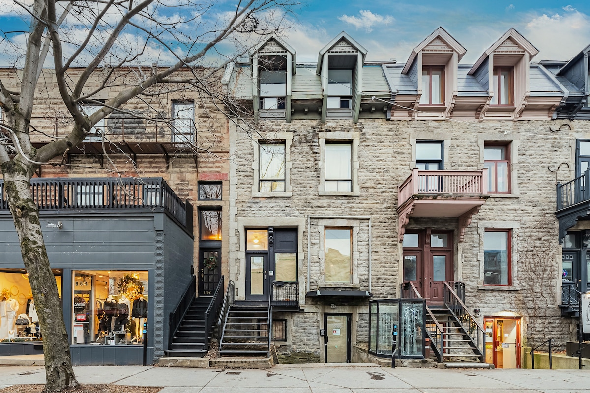 The image showcases a classic stone building in a vibrant urban setting. The exterior features multiple levels with dormer windows and balconies. A tree is visible in the foreground, while shops and a staircase lead up to the entrance, highlighting the architectural charm of the neighborhood.