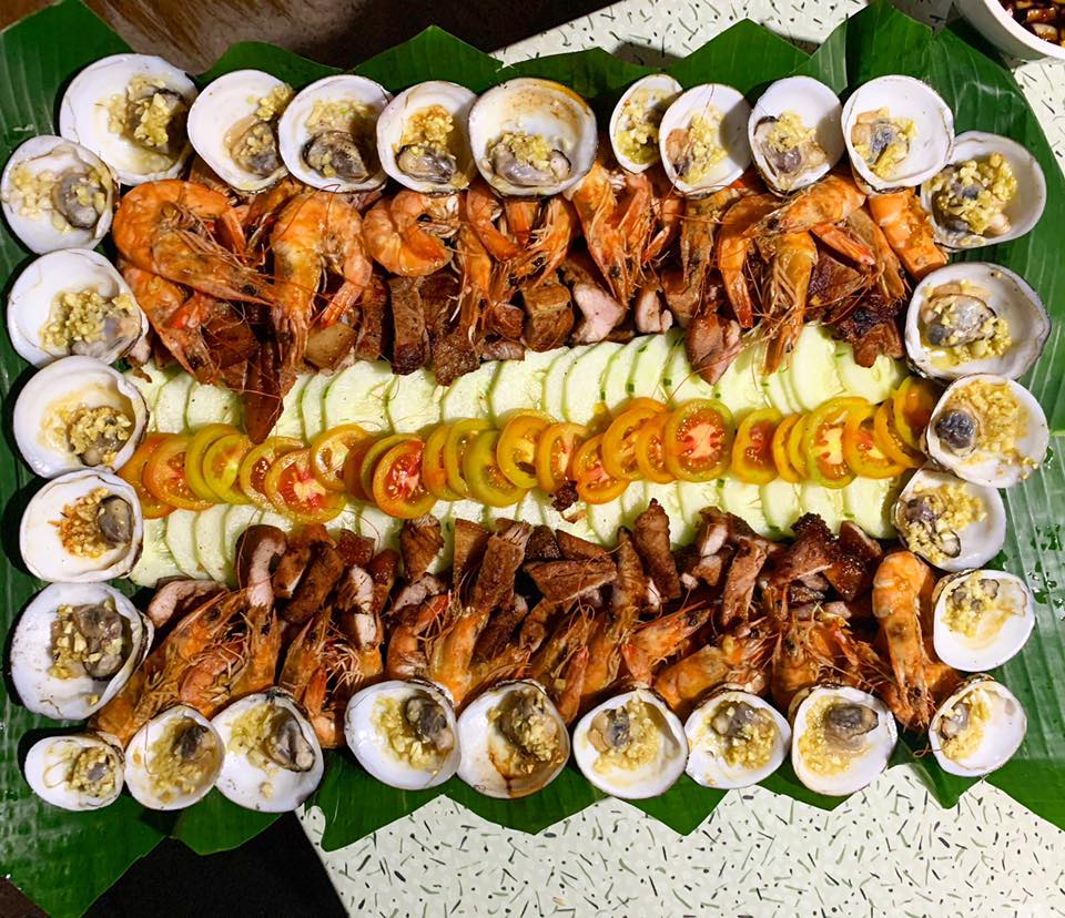 An assortment of grilled shrimp and clams is displayed on a large platter lined with green banana leaves. Slices of cucumber and tomato are arranged between the seafood, adding freshness and color to the presentation.