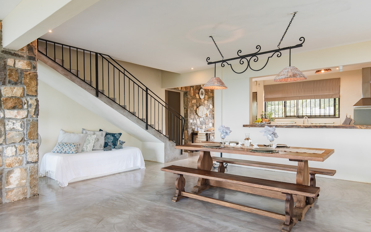 An open living area features a wooden dining table accompanied by decorative table settings. A staircase with a wrought iron railing leads to the upper level. Natural light illuminates the space, complementing the stone wall accents and the adjacent kitchen area laid with modern fixtures.