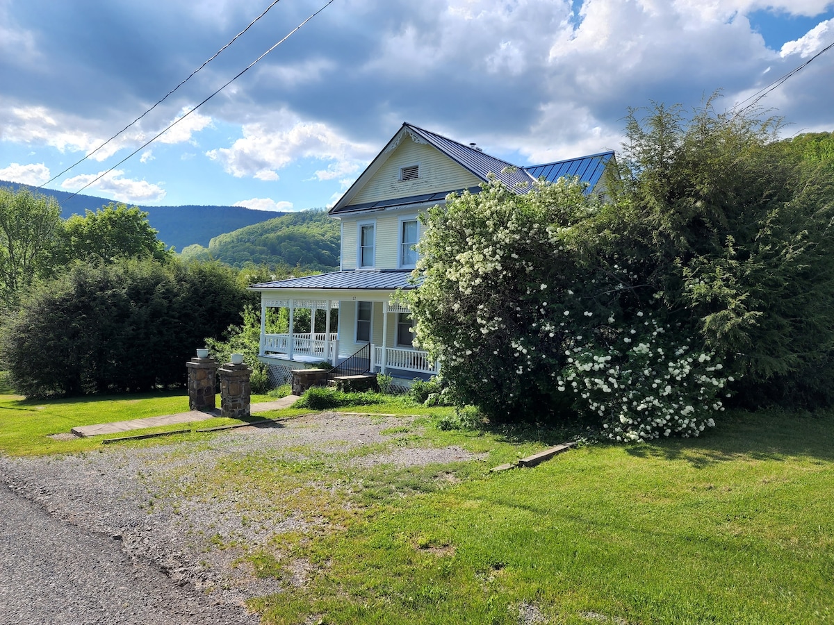 The restored Victorian house is framed by lush greenery and flowering bushes, set against a backdrop of rolling hills. A wrap-around porch is visible, offering a welcoming space. A gravel pathway leads to the entrance, and expansive grassy areas surround the property.