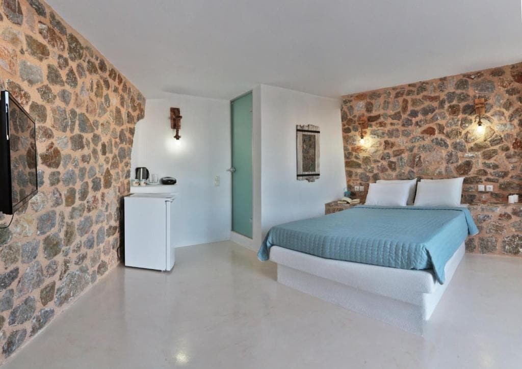 A spacious bedroom features a large double bed with a light blue coverlet against a backdrop of natural stone walls. A small kitchenette area is visible, equipped with a refrigerator and kettle. Soft lighting enhances the neutral tones throughout the room.